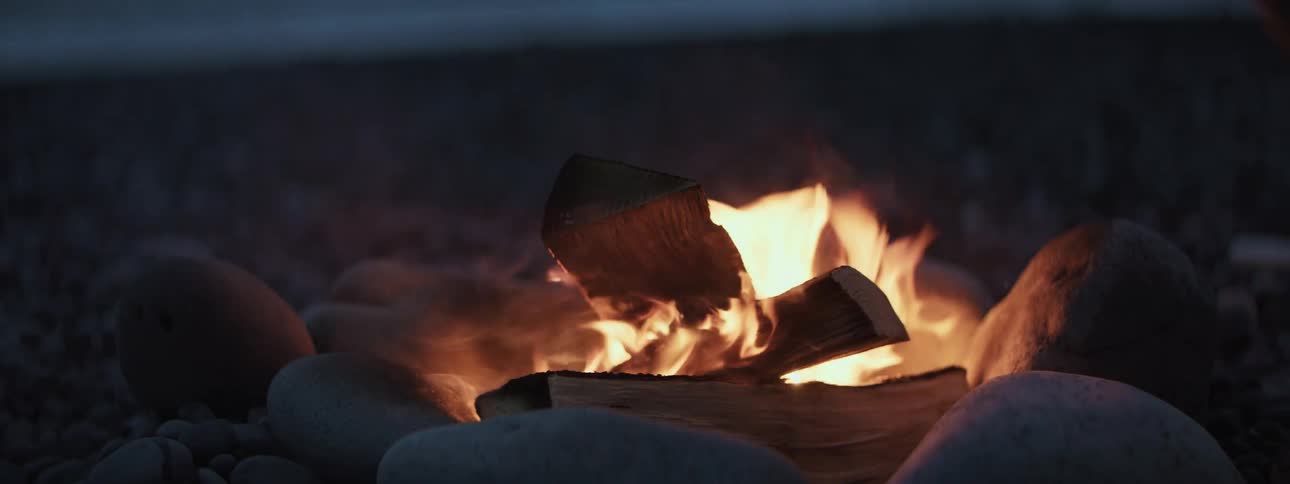 A cozy campfire burns brightly on a rocky shore at night, with sparks flying from the flames