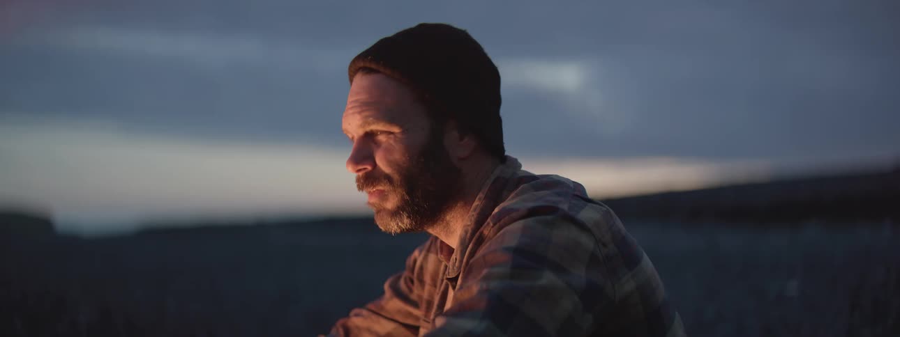 Close-up of a contemplative man warming himself by a crackling campfire on a rocky beach at dusk