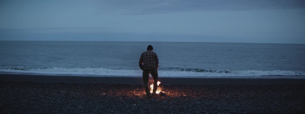 A solitary man tends to a small campfire on a rocky beach while watching the ocean waves at twilight