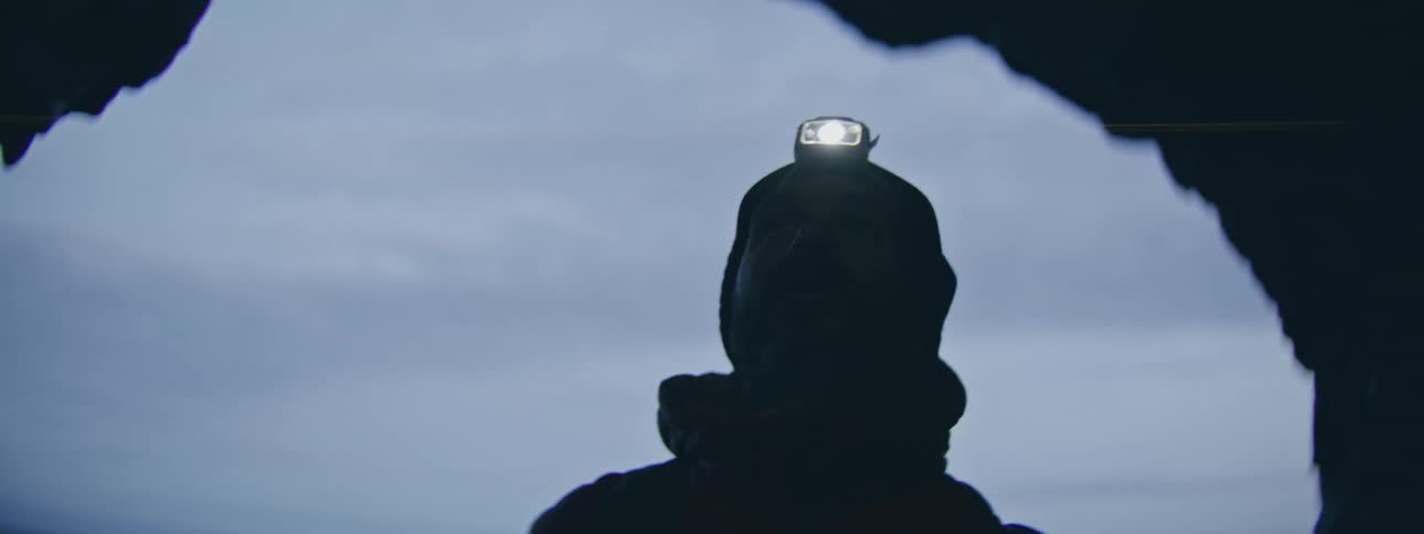 A silhouetted man turns on his headlamp inside a dark cave, creating a beautiful lens flare