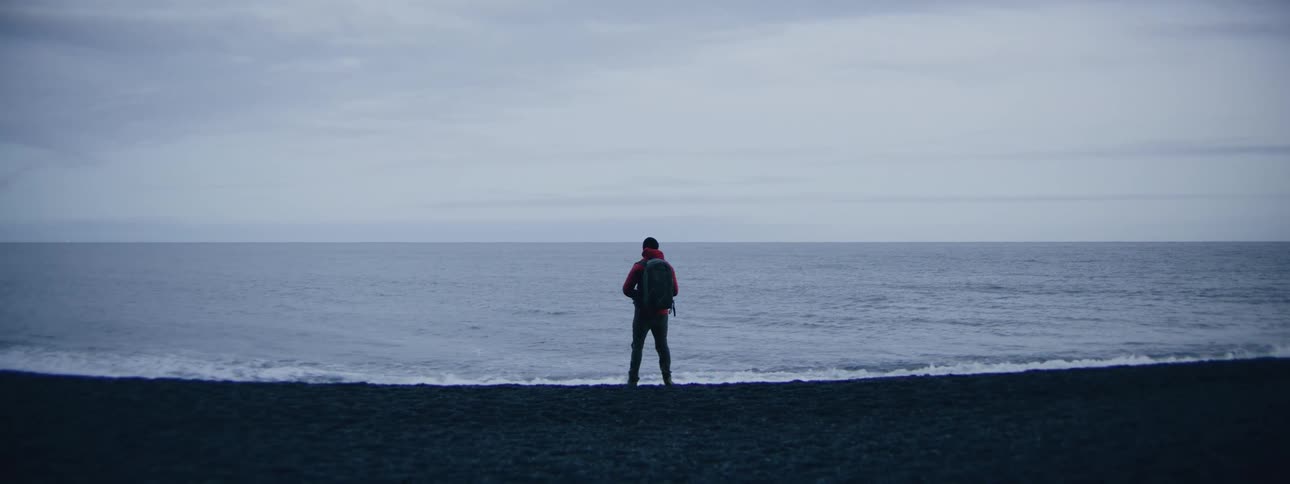 A photographer in a red jacket walks along a desolate black sand beach by the ocean on a moody day
