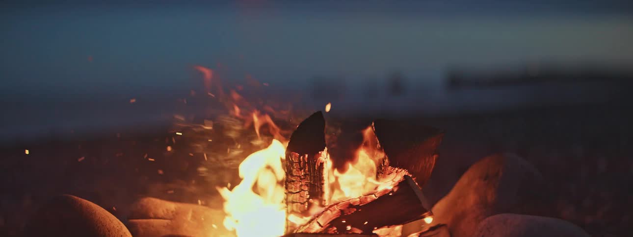 A slow-motion close-up of a crackling campfire on a beach at night, with sparks flying from the glowing embers