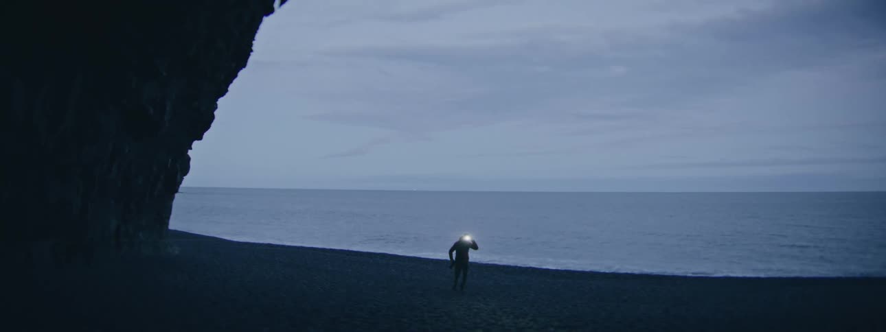 A solitary adventurer with a backpack turns on a headlamp and walks along a dark sand beach at night