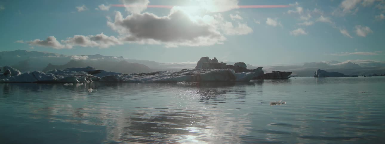 A serene wide shot of a glacial lagoon with icebergs floating in the calm water as the bright sun shines overhead