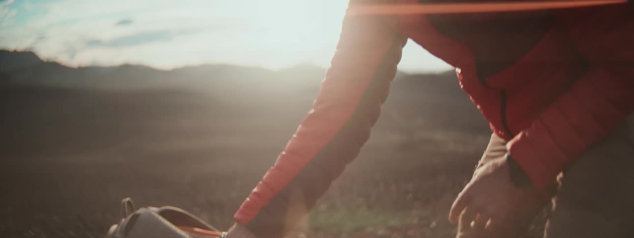 An adventurer in a red puffer jacket unpacks his bag on a windy mountain peak at sunrise