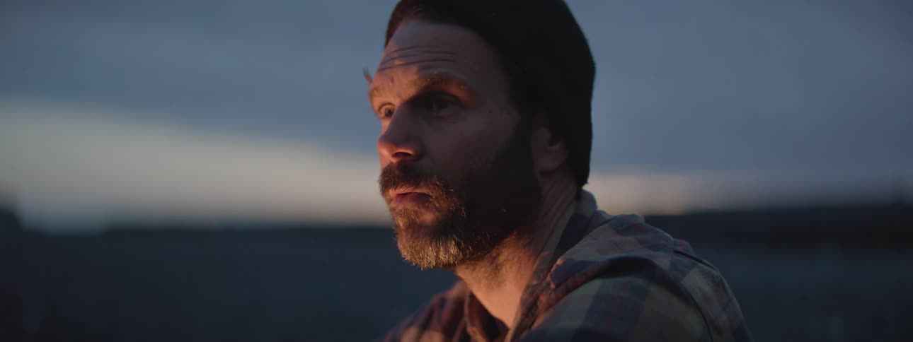 A contemplative bearded man in a beanie sits by a campfire at dusk, his face illuminated by the warm, flickering light