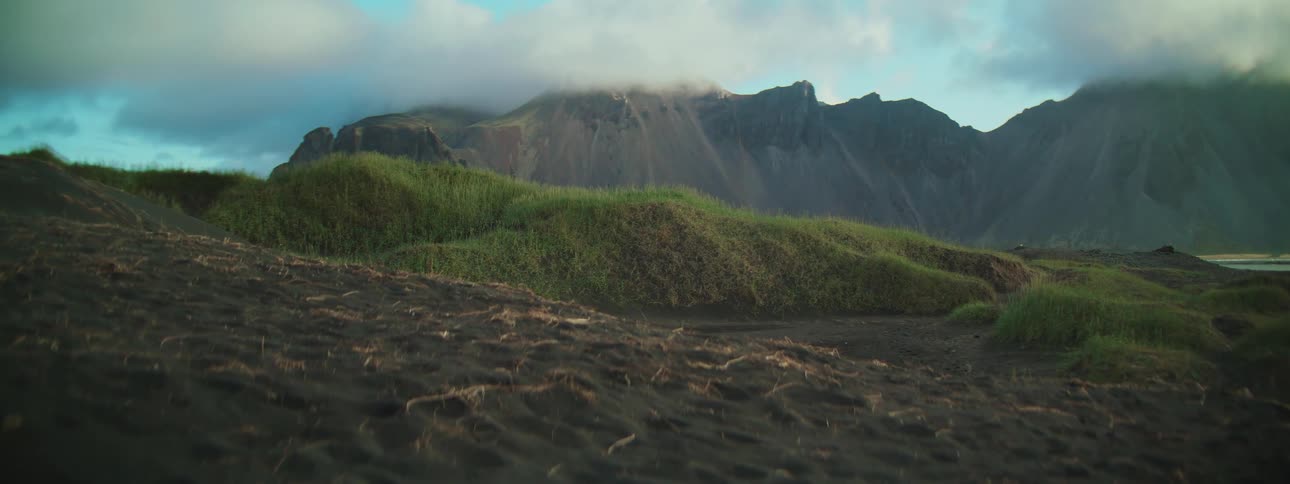 Low clouds drift over the rugged peaks of Vestrahorn mountain above vibrant green grass dunes and volcanic black sand