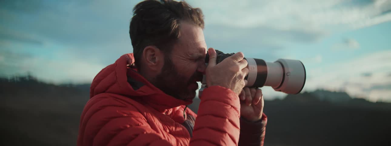 A bearded male photographer in a red jacket brings his camera with a telephoto lens to his eye to take a picture outdoors