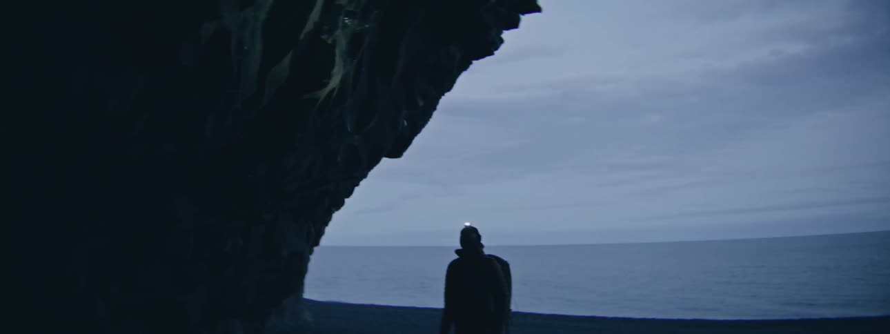 An adventurer wearing a headlamp explores a dark, rocky seaside cave on a black sand beach at twilight