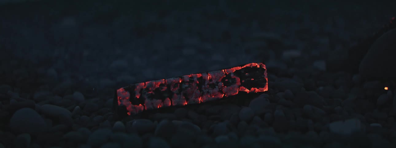 A single smoldering log glows with hot red embers on a pebble beach at night