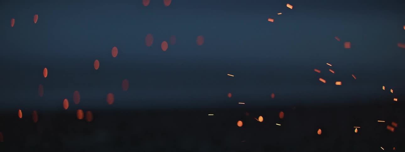 Out-of-focus shot of glowing orange sparks and embers floating up from a bonfire in slow motion at night