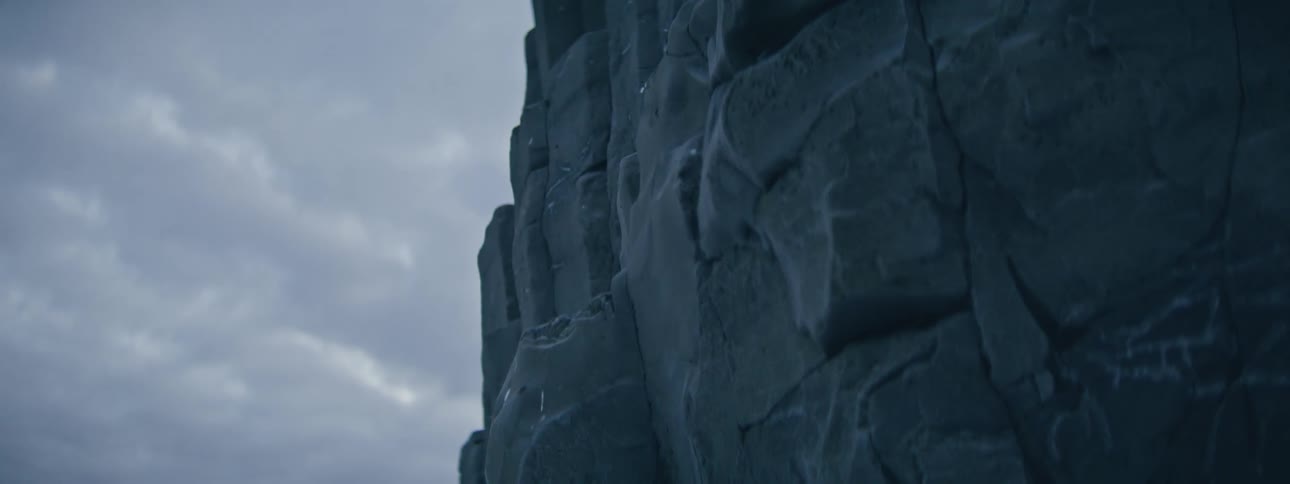 A dramatic close-up reveals the icy texture of a dark, jagged rock formation under an overcast sky