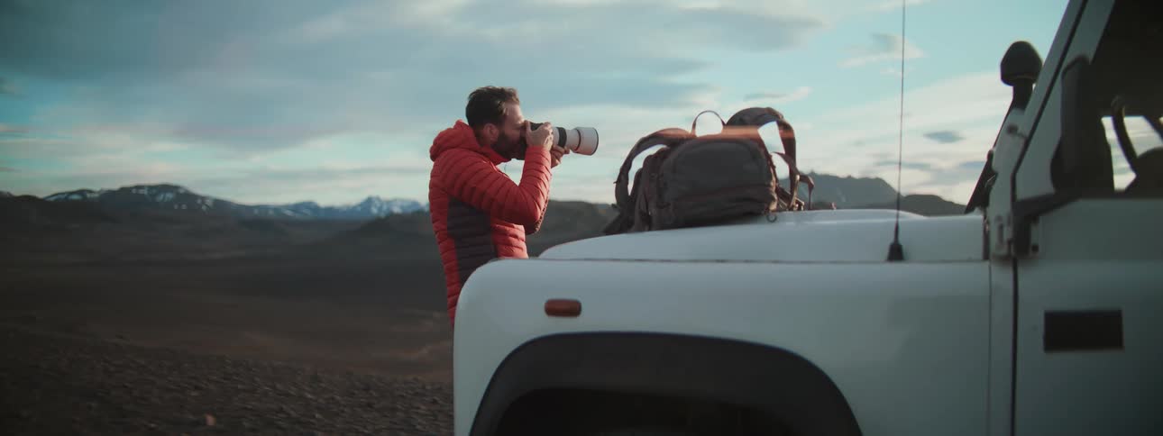 An adventurous photographer in a red jacket captures the epic mountain landscape with a large camera lens next to his truck
