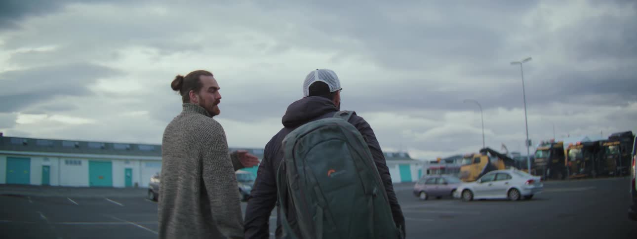 Two male friends with backpacks walk and talk through an industrial parking lot on an overcast day