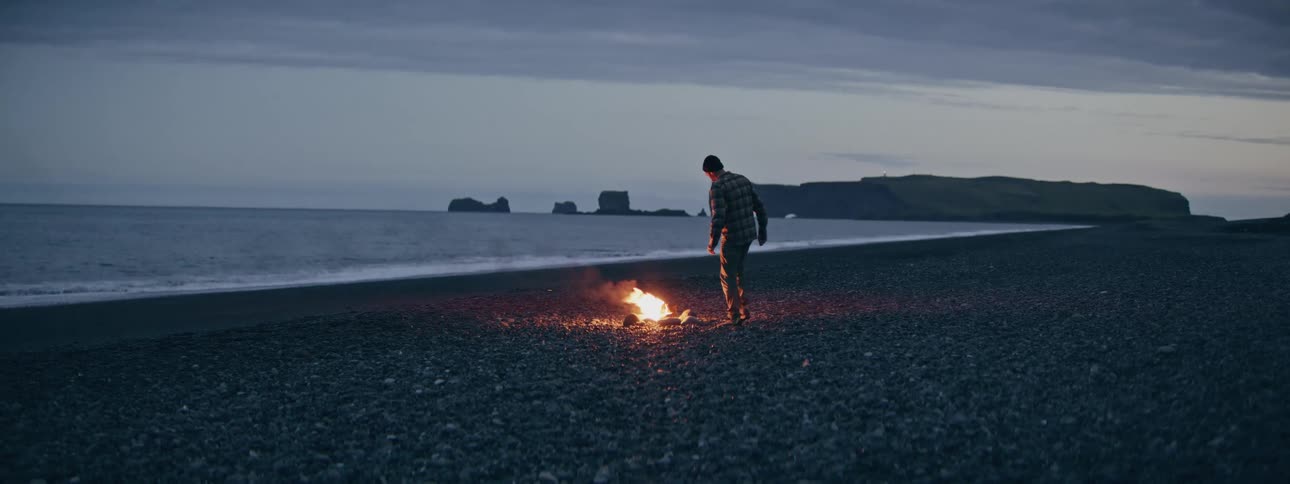 A man walks towards a bonfire on a dark, pebbly beach at dusk and crouches to warm himself by the flames