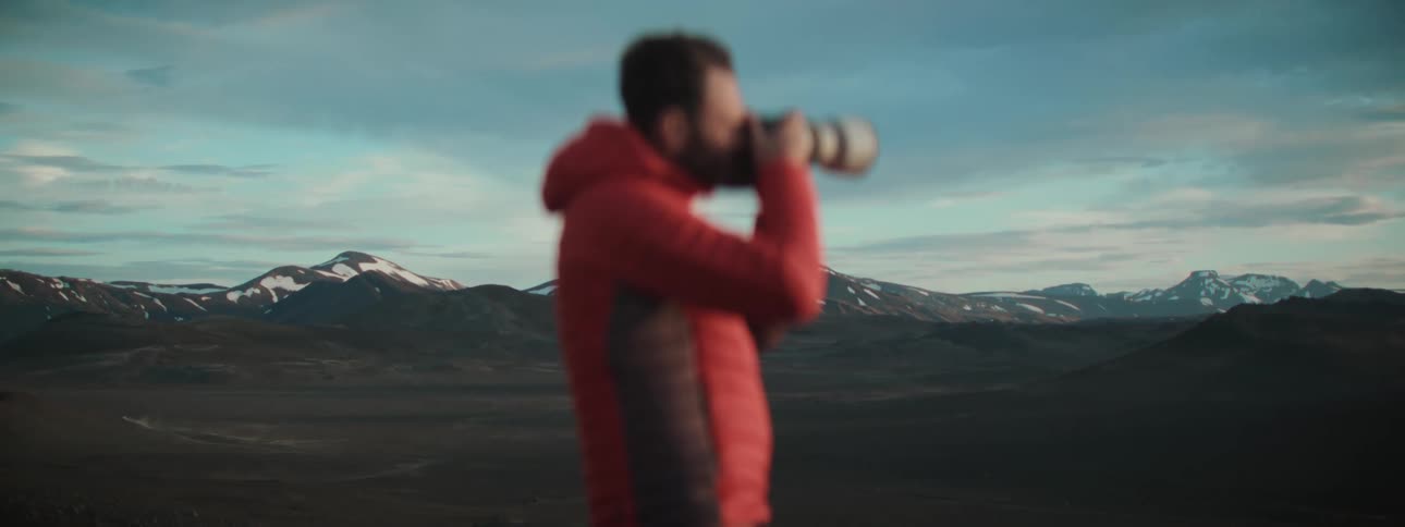 An out-of-focus photographer in a red jacket uses a telephoto lens to capture the vast, snowy mountain landscape at dusk