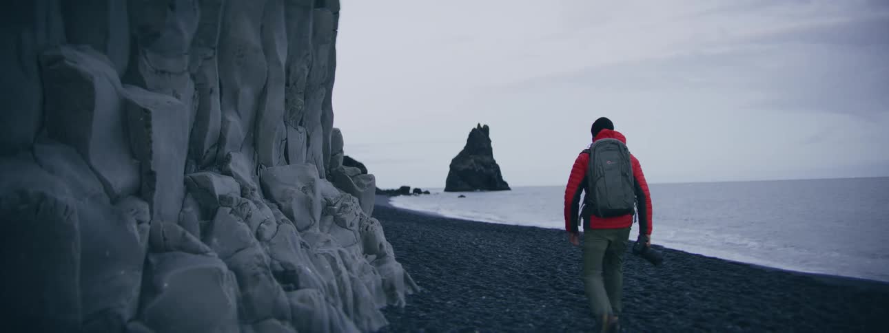 An adventurous photographer in a red jacket walks along a stunning black sand beach in Iceland