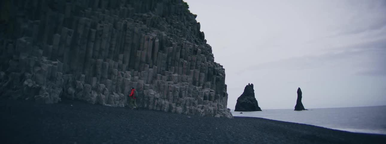 A hiker walks along a black sand beach with dramatic basalt column cliffs in Iceland