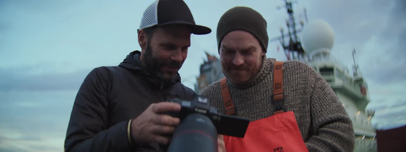 A photographer and a fisherman share a smile as they look at photos together on a camera screen while on the deck of a fishing boat