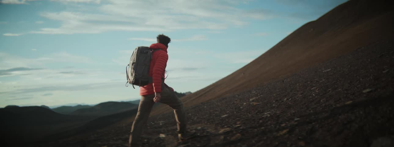 A determined male hiker ascends a steep, barren mountainside in slow motion during golden hour