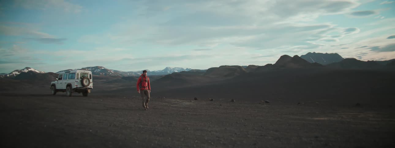 A male adventurer in a red jacket walks away from his white off-road vehicle to explore a vast and remote volcanic landscape in slow motion