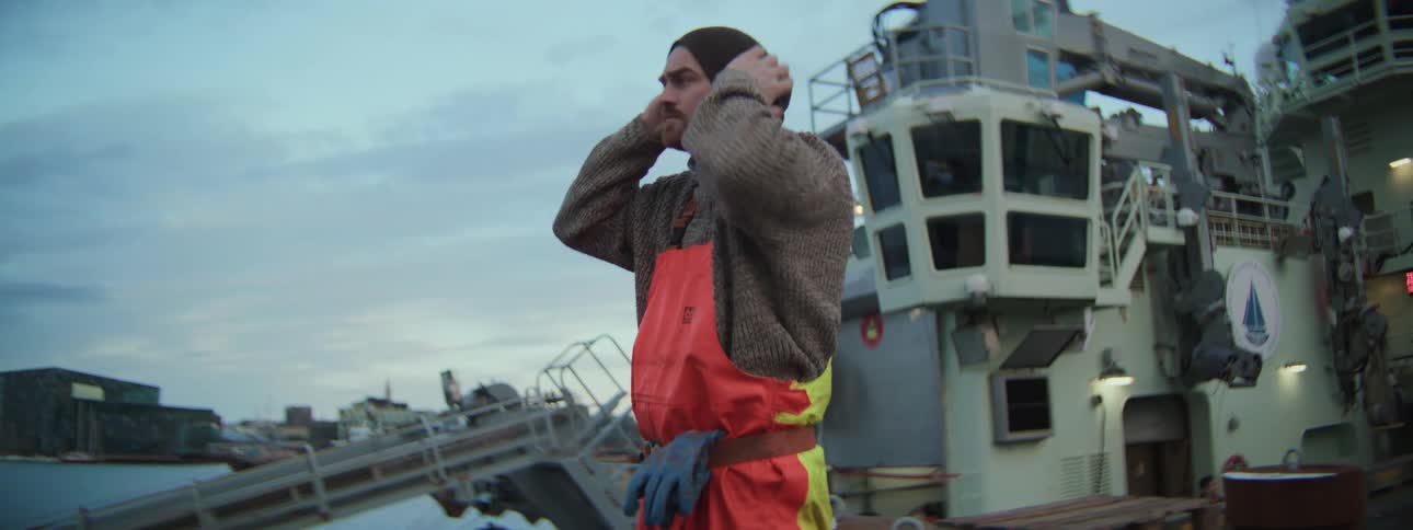 A fisherman in a thick sweater and orange overalls puts on his beanie and rolls up his sleeves on the dock