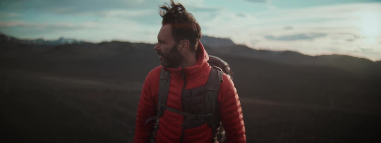 A male hiker in a red jacket treks through a vast, windy volcanic landscape in dramatic slow motion