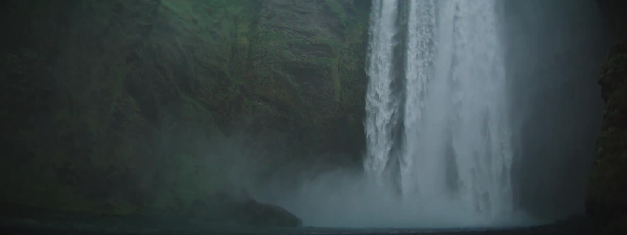 A powerful waterfall cascades over a lush green cliff face as the camera tilts down into the mist below