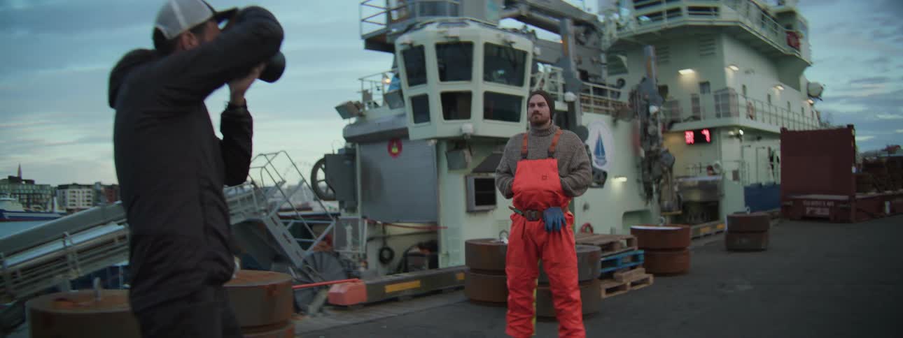A photographer takes a portrait of a rugged fisherman in overalls standing on a commercial dock in front of a large industrial ship