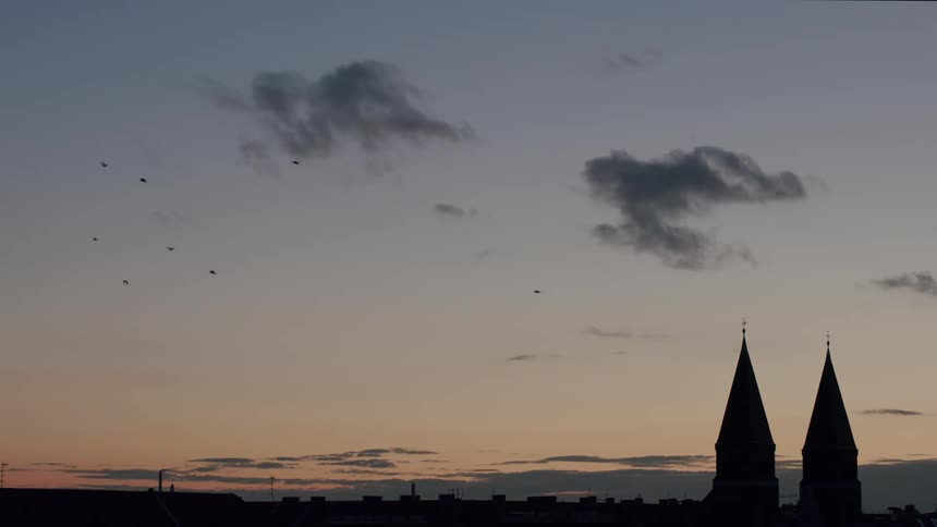A flock of birds flies across a beautiful sunset sky above the silhouetted spires of a city church