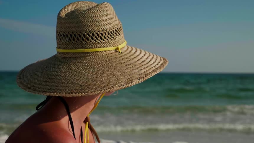 A person wearing a straw hat stares out toward the horizon at a beach