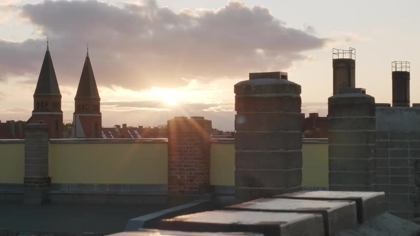 The setting sun casts a warm glow over a European city's rooftops, with church steeples and chimneys silhouetted against the cloudy sky