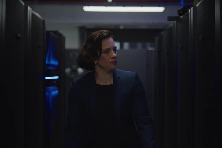 A serious female IT professional in a business suit walks through a dark server room while inspecting the server racks