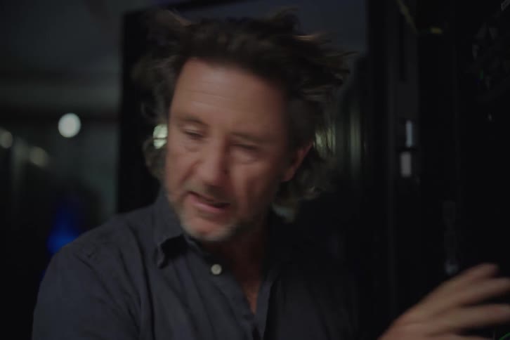 A flustered man in a dark server room has his hair blown back by a fan while trying to solve a technical issue