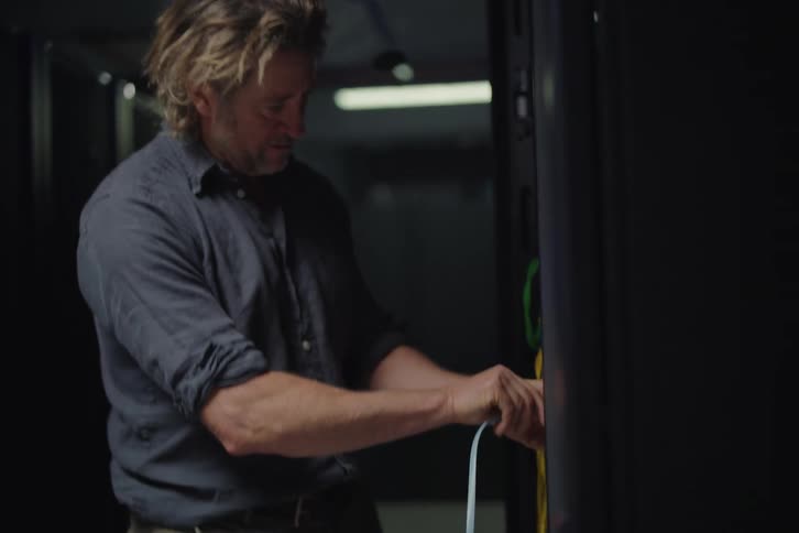 An IT technician discusses a network issue over a laptop before plugging an ethernet cable into a server rack in a dark data center