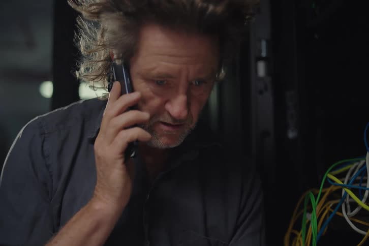A stressed and confused IT support engineer with hair blowing in the wind talks on the phone while trying to fix a messy server rack