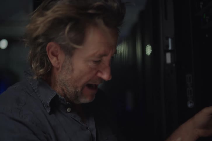A frantic IT technician troubleshoots a network issue in a server room as a fan blows his hair around