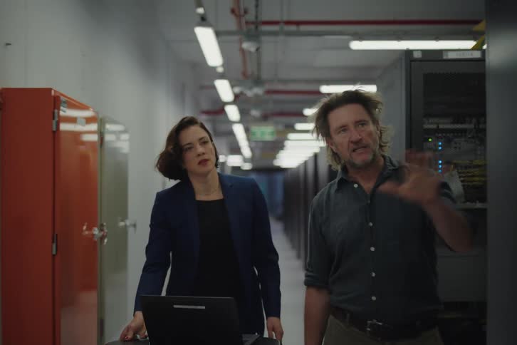 An IT technician passionately explains the server room's functions to his female colleague as they walk down the aisle of a modern data center