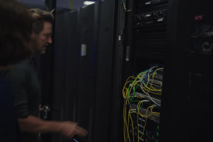 A male IT engineer walks with a female manager through a server room, stopping to inspect and explain the computer network equipment