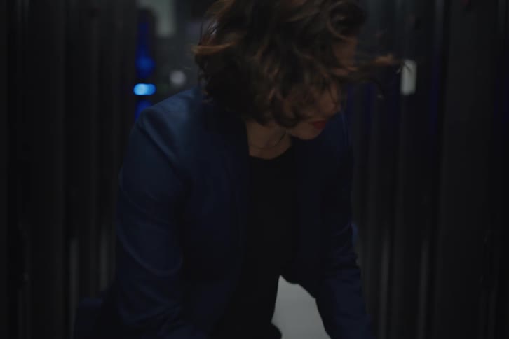 A focused female IT technician works on her laptop in the cool, windy aisle of a data center