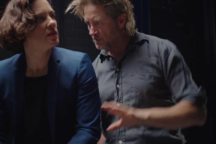 A male and female IT professional collaborate to solve a problem in a server room, with the man explaining while the woman works on a laptop