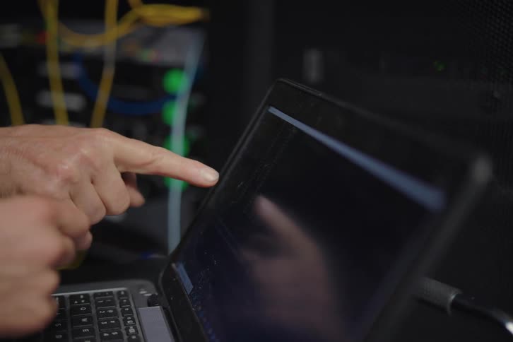 A shallow depth-of-field close-up shot of two IT technicians' hands pointing at server cables before rack focusing to them diagnosing the issue on a laptop