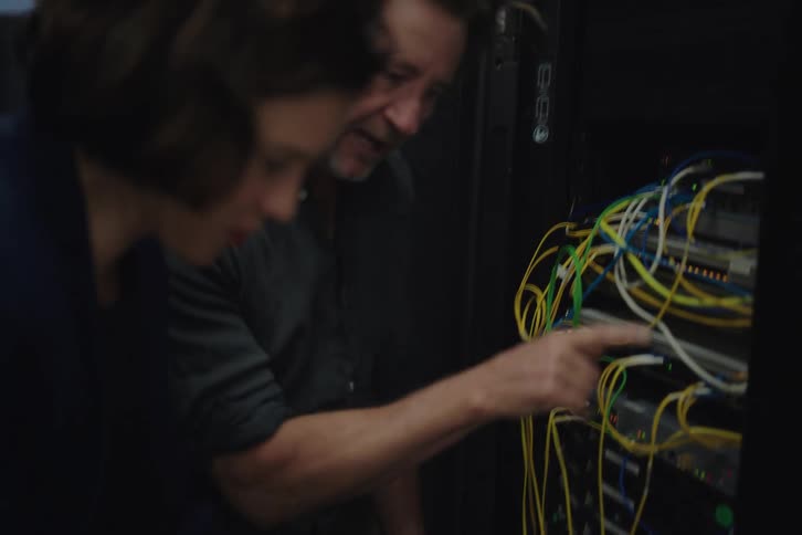 A male and female technician collaborate in a dimly lit server room, troubleshooting network issues on a laptop