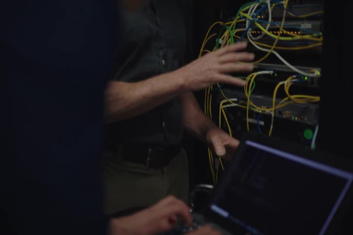 A male IT technician explains an issue with a server rack while a female colleague works on her laptop in a data center