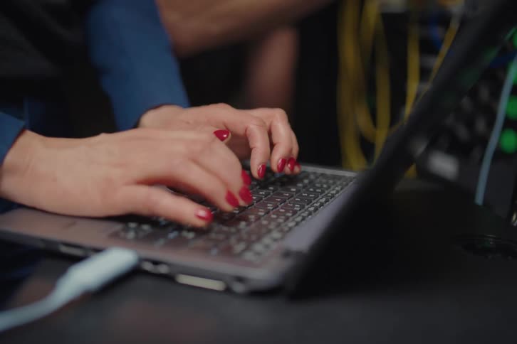 A close-up of a female IT engineer's hands typing on a laptop keyboard as her male colleague assists in a server room