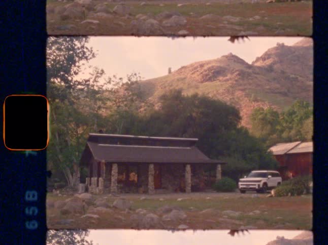 Vintage Super 8 film captures a rustic stone cabin and a parked SUV nestled in a serene, hilly landscape