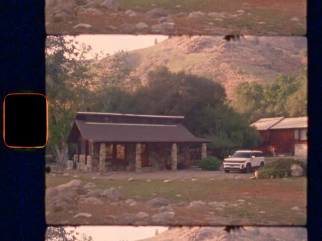 A Super 8 film shot tilts down from a grassy mountain to a rustic stone cabin nestled in the trees