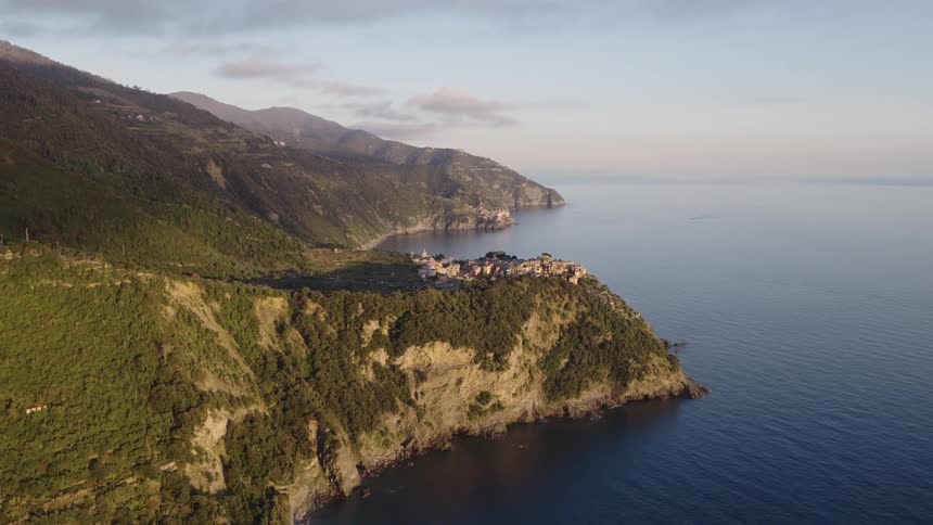 A stunning aerial view of the historic village of Corniglia in Cinque Terre, perched on a cliffside overlooking the calm sea at sunrise