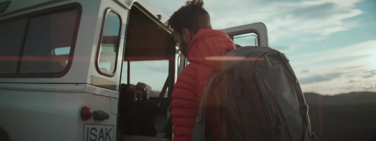 A man in a red jacket grabs his backpack from an off-road vehicle and begins a solo hike through a vast, remote landscape at sunrise
