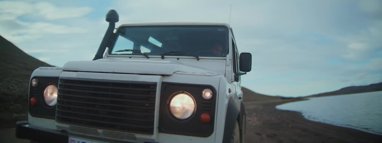 A man in a red jacket drives a white off-road vehicle along a lakeside dirt road, stopping to get out and admire the rugged landscape